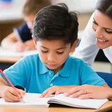 Criança estudando em carteira escolar com a professora ajudando no caderno